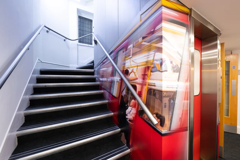 A set of stairs at Broadway House next to a lift. The adjacent wall has a decorative painting of a London Underground train.