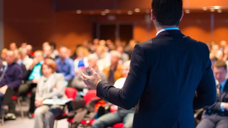 Person in the foreground presenting in front of a crowd of seated people.